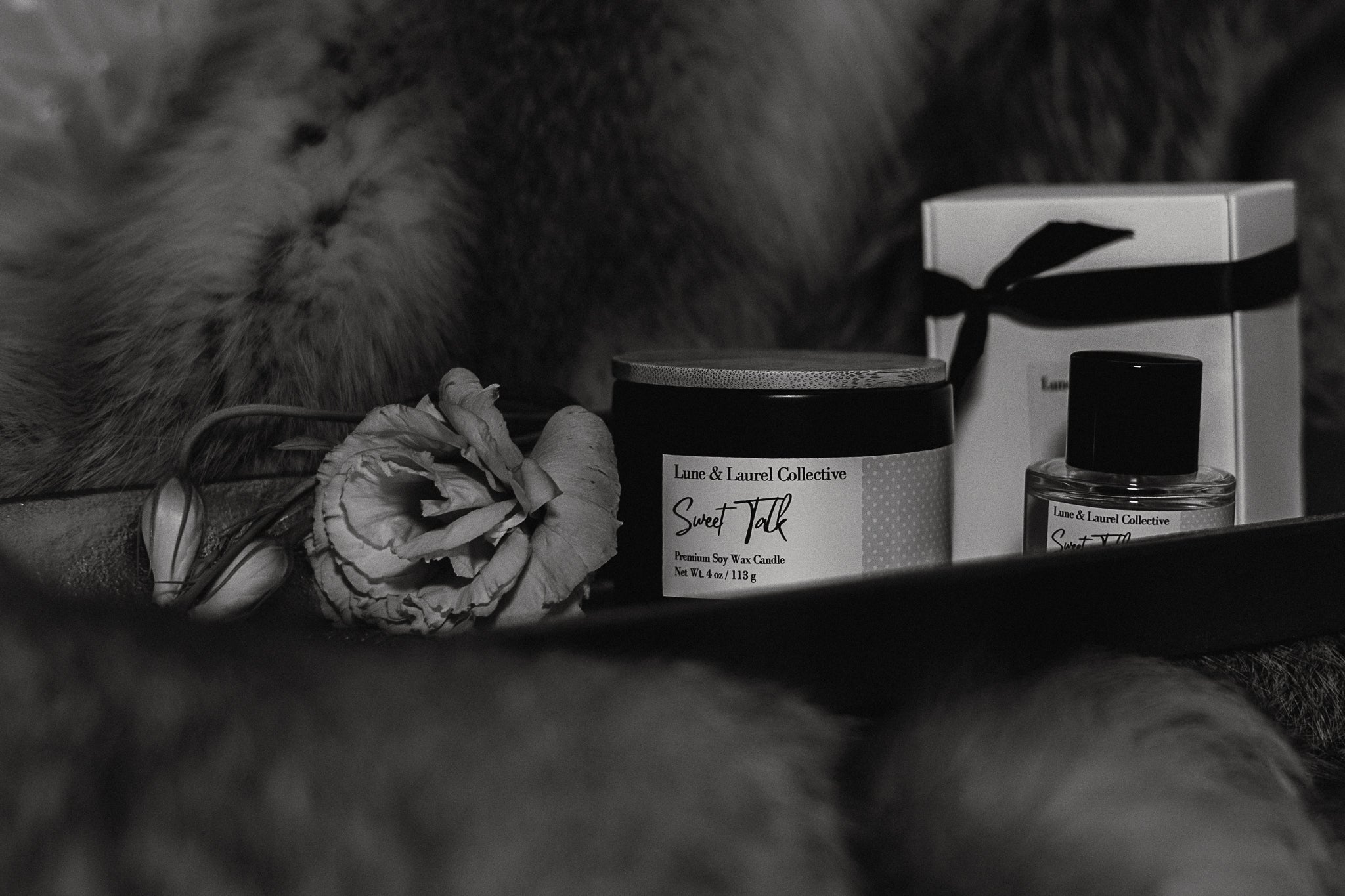 A black and white photograph showcasing the "Sweet Talk" scent from the Signature Collection by Lune & Laurel Collective. In the center sits a dark 4 oz premium soy wax candle with a bamboo lid. To the right is a clear 30ml perfume bottle beside its white gift box tied with a dark ribbon. To the left rests a delicate open flower on a stem, all arranged on a gold tray against a soft fur backdrop.
